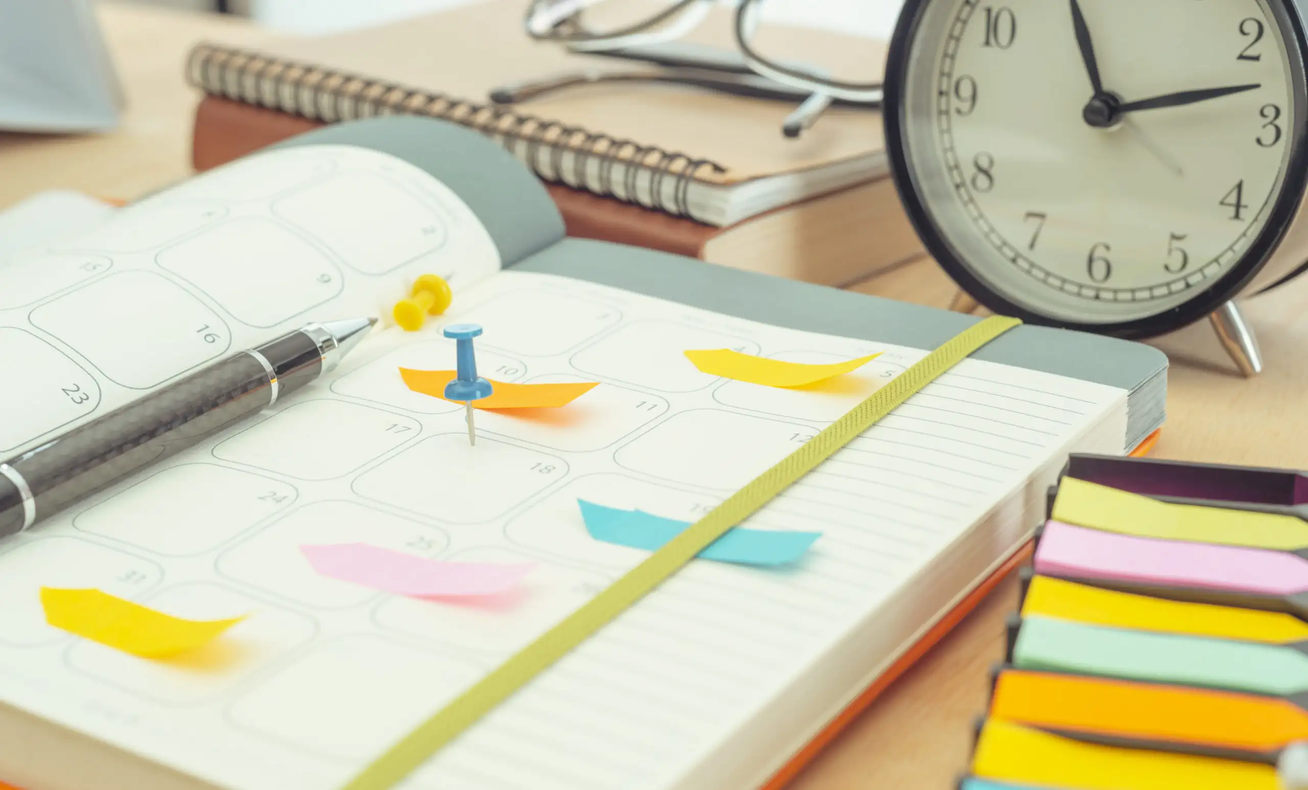 Daily planner open on a messy desk with pens, pins, glasses, sticky notes, and a clock