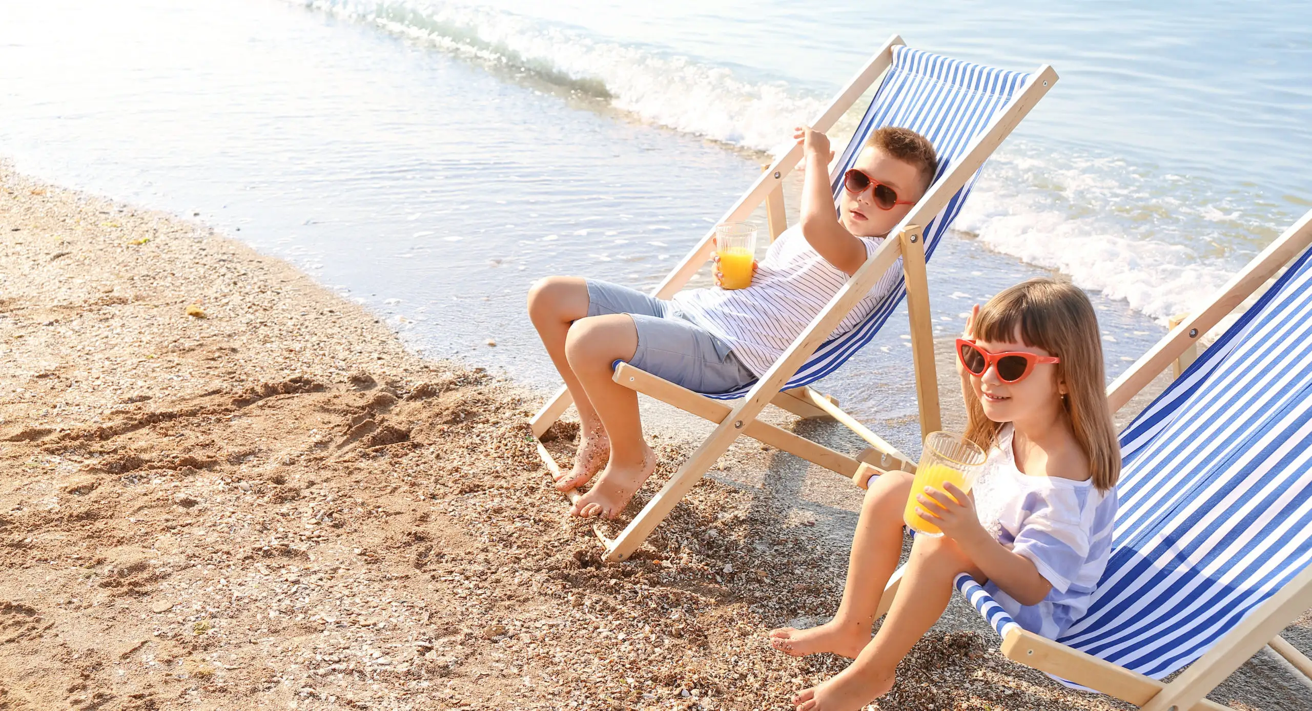 Two children drinking juice on the beach in beach chairs