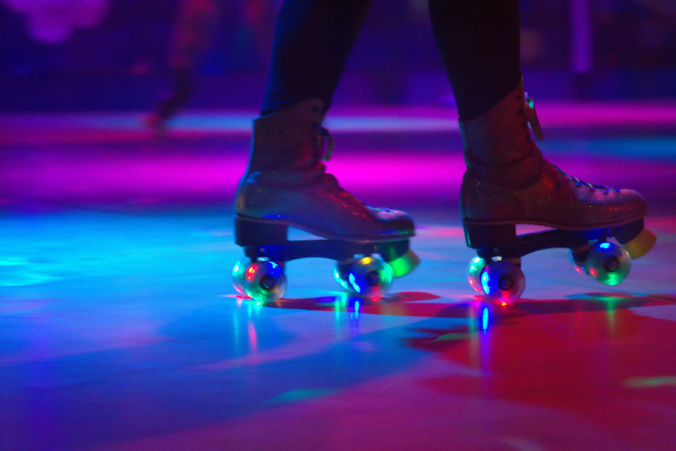 A person from the shins down, wearing a pair of roller skates in a dark roller rink with pink and purple lights