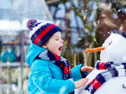 little kid boy making a snowman and eating carrot. child playing and having fun with snow on cold day.; Courtesy of Romrodphoto/Shutterstock