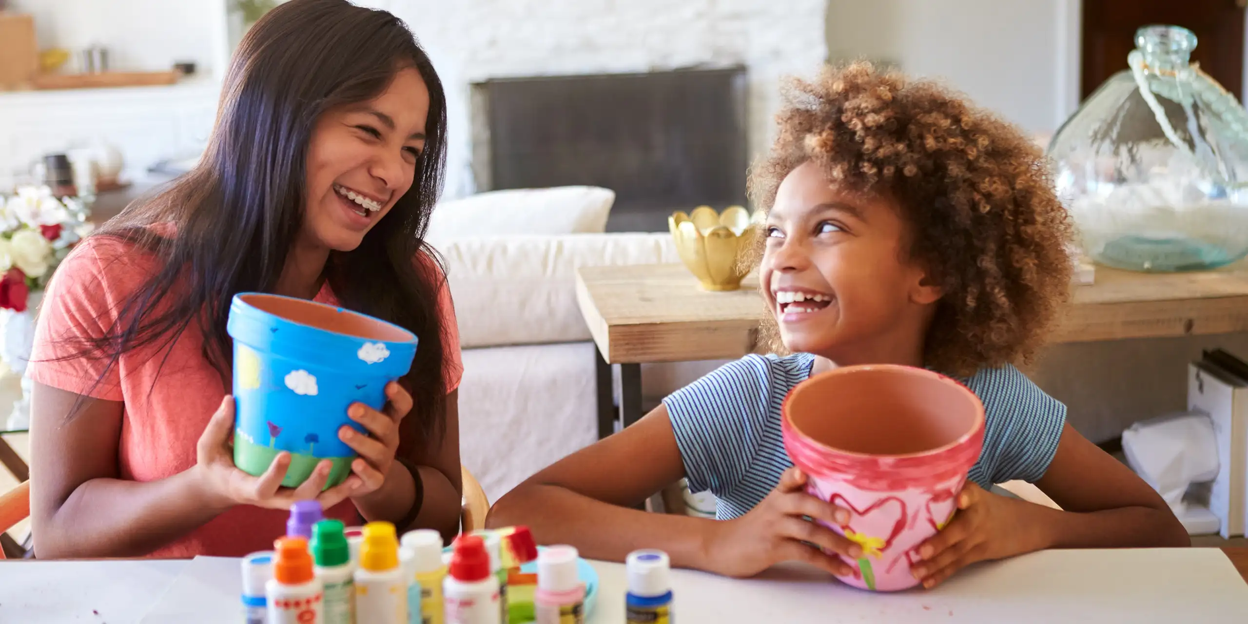 Happy pre-teen girl and her older girlfriend holding plant pots that theyâ€™ve decorated with paints at home, smiling at each other